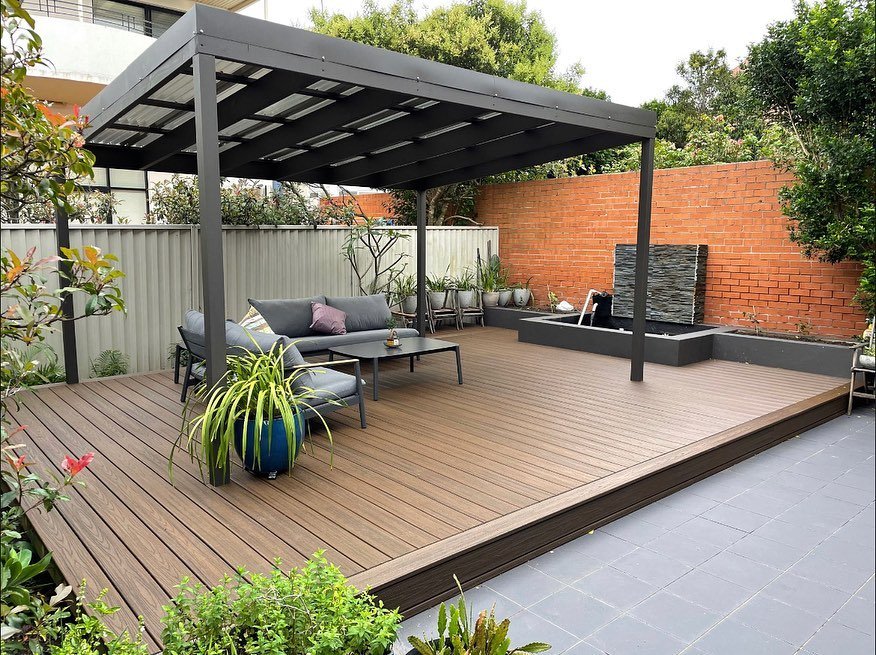 Dark aluminium pergola with translucent polycarbonate roof over a composite deck and outdoor lounge